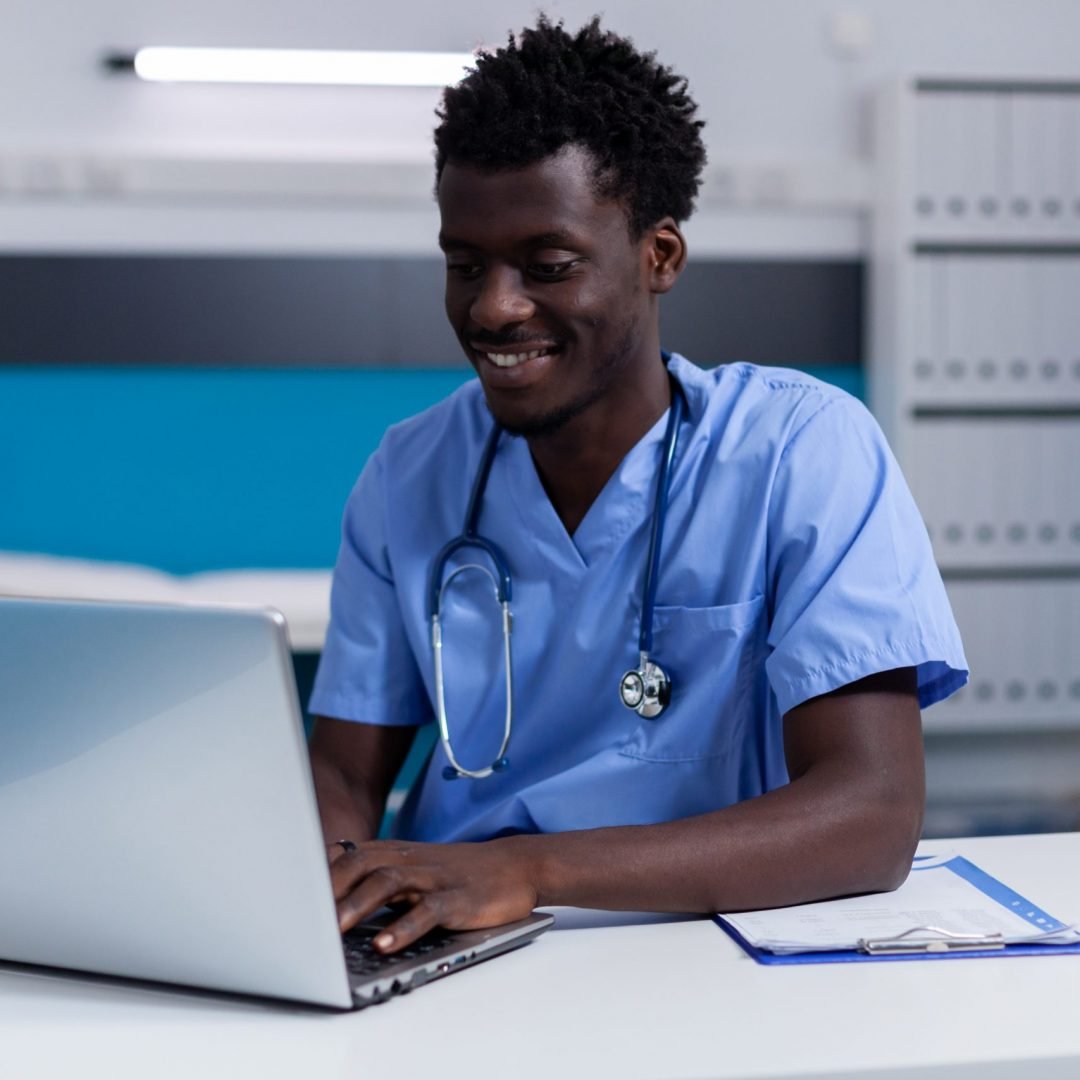 Black young man working as nurse at medical clinic wearing uniform and stethoscope. African american person sitting at desk while using modern laptop, typing on keyboard in cabinet