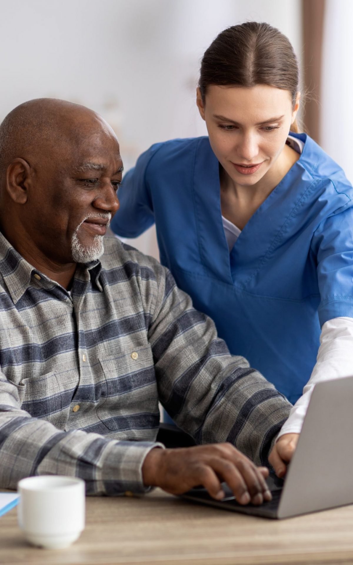 Friendly young woman nurse helping black senior man in weheelchair using laptop, smiling elderly african american male patient surfing on Internet at nursing home, copy space